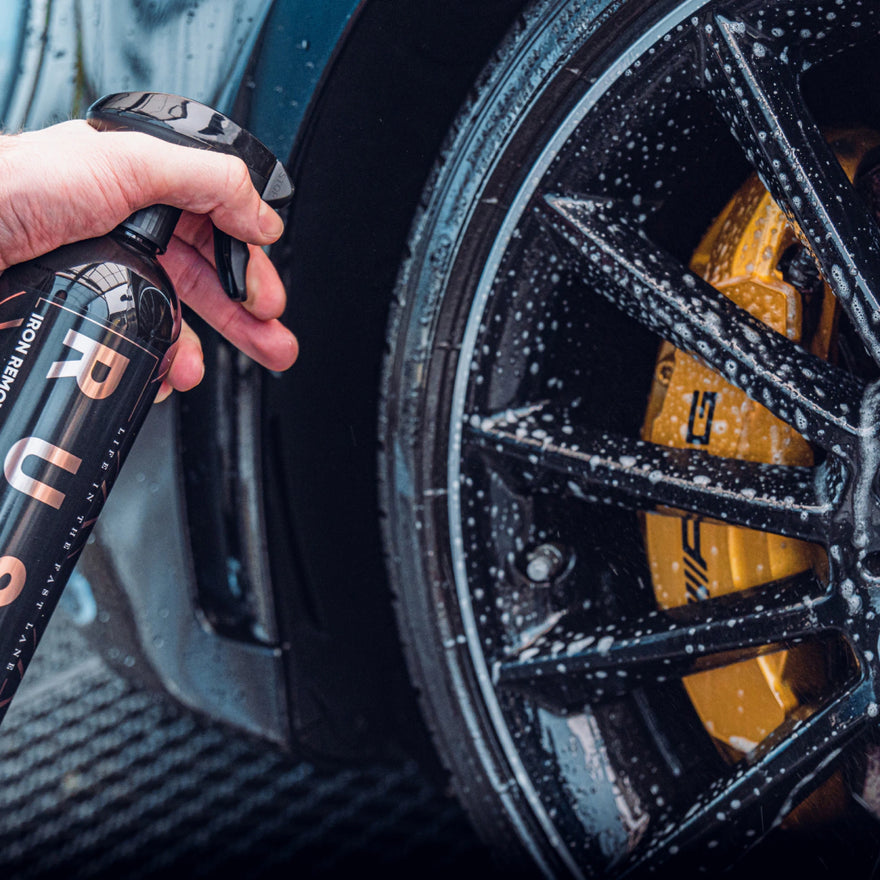 A hand sprays Iron Remover onto the soapy black wheel and yellow brake caliper, using it to clean the rim as part of a vliegroest verwijderen routine.