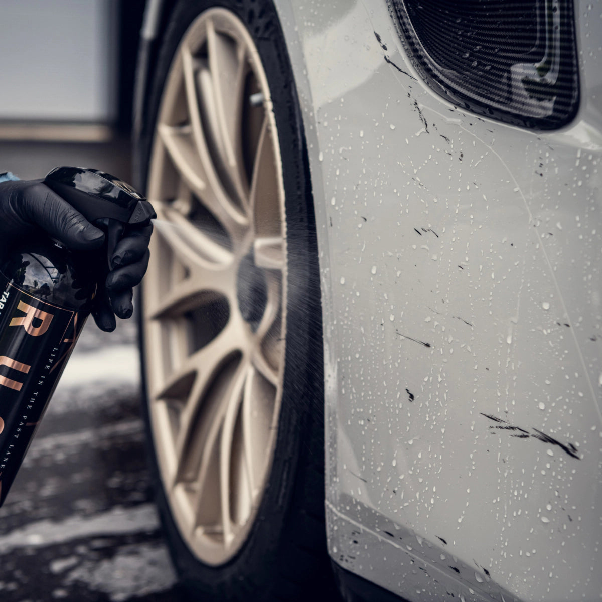 Een gehandschoende hand spuit teerverwijderaar op een vuil autowiel en pakt vuil en waterdruppels op de witte carrosserie van de auto aan.