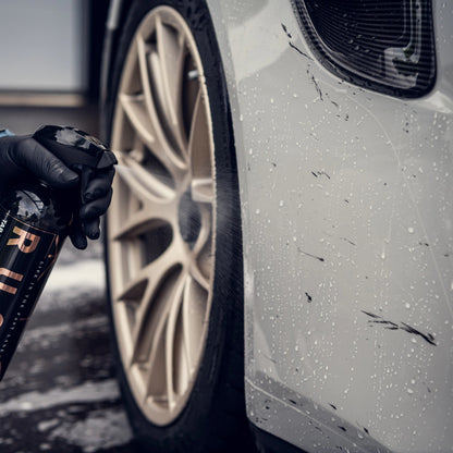Een gehandschoende hand spuit teerverwijderaar op een vuil autowiel en pakt vuil en waterdruppels op de witte carrosserie van de auto aan.