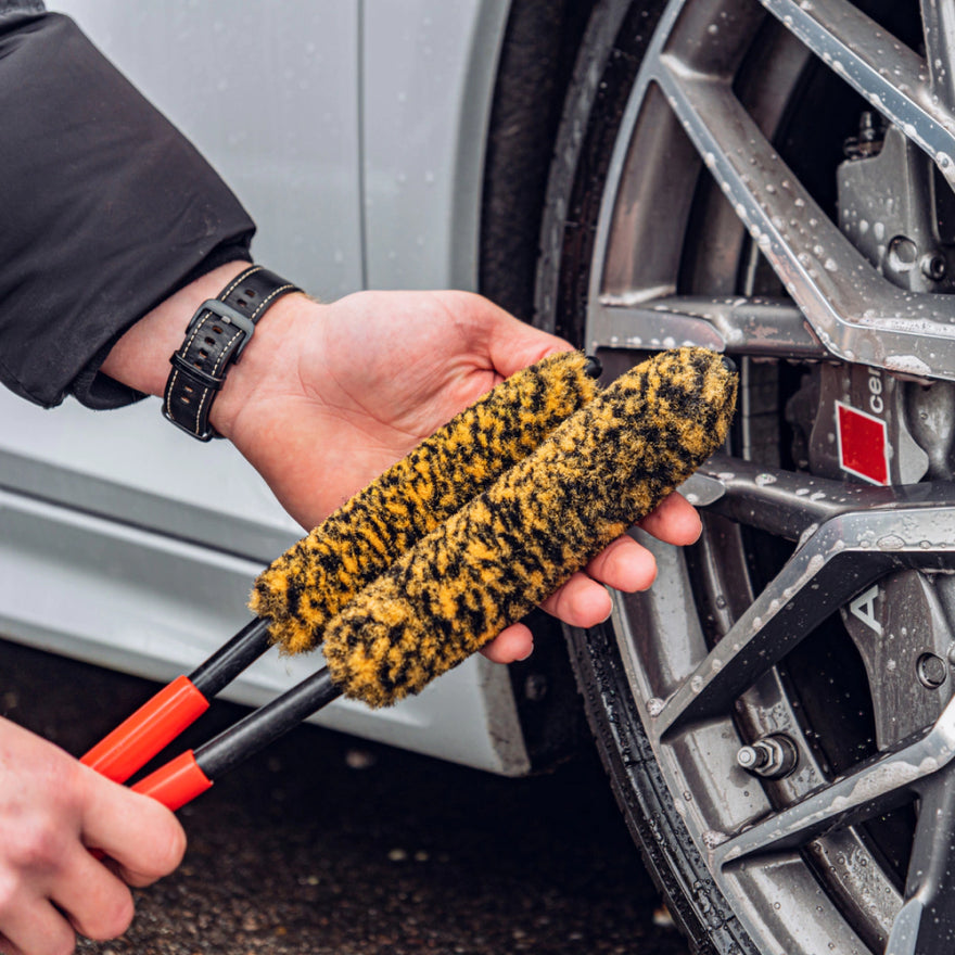 Someone holds two yellow and black Wheel Woolie Set brushes near a car's silver alloy wheel, preparing to clean the spokes. The Wheel Woolie Set ensures effective cleaning and easily reaches every corner during velgen reinigen.