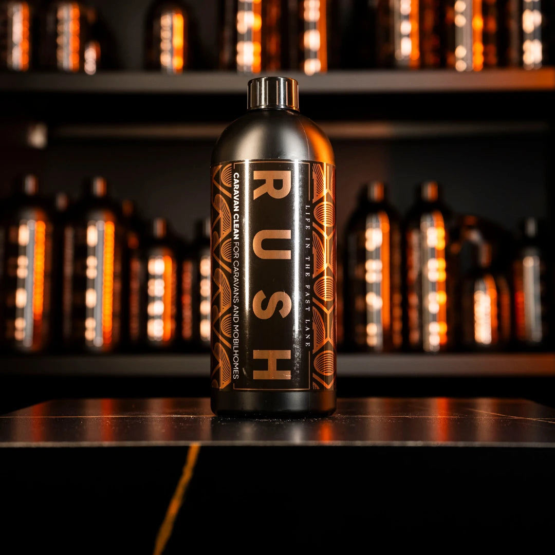 Een zwarte fles met het label RUSH, perfect als caravan reiniger, staat op een donker oppervlak met vage flessen op de achtergrond.