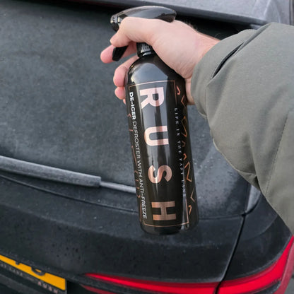 A hand in a grey sleeve holds a black De-Icer spray bottle labeled RUSH DE-ICER WITH ANTI-FREEZE in front of a frosted car rear window and trunk.