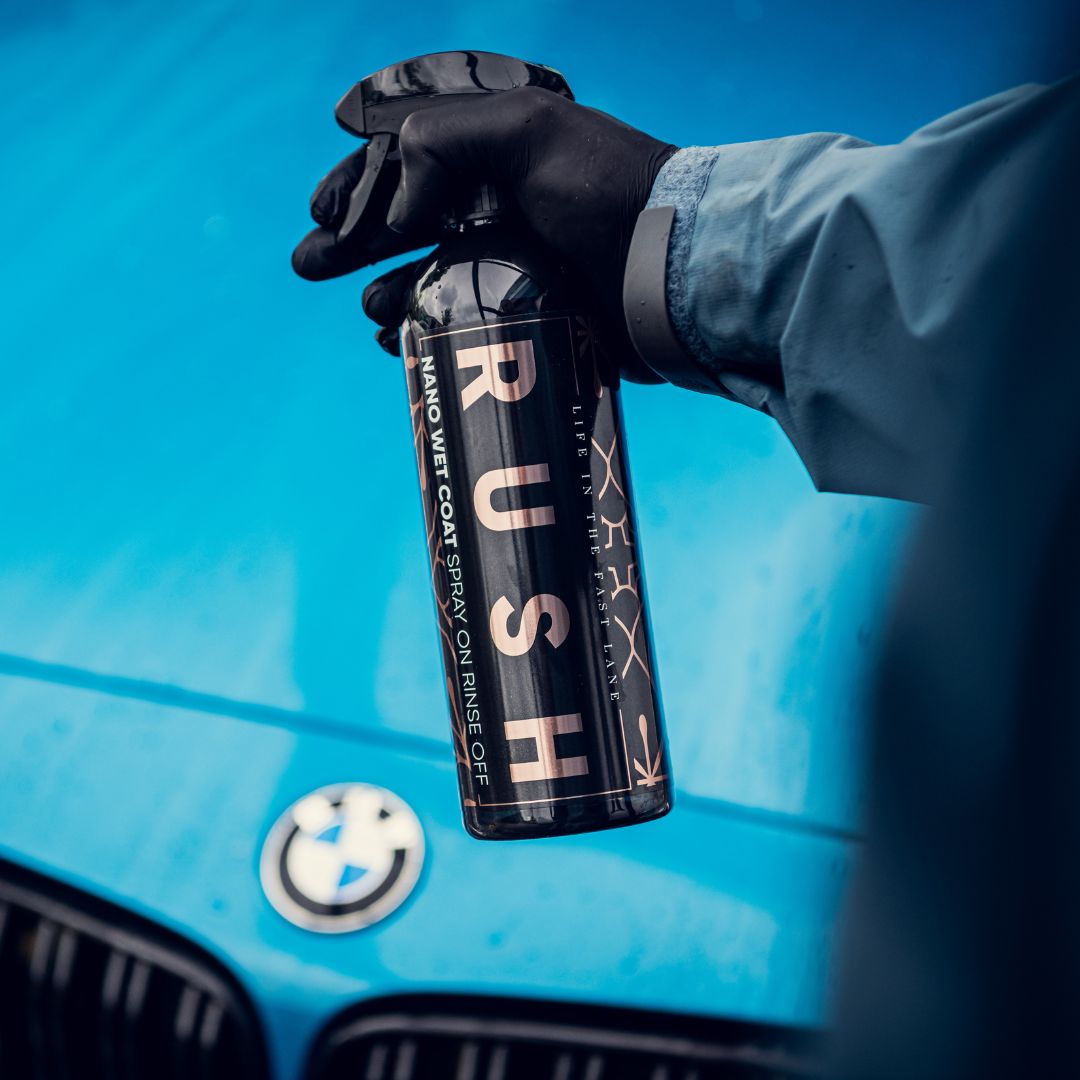 A gloved hand holds a bottle labeled RUSH Nano Wet Coat in front of a blue BMW hood with the emblem—an essential part of the Detailing Package PLUS | 23-delig for achieving a flawless finish.