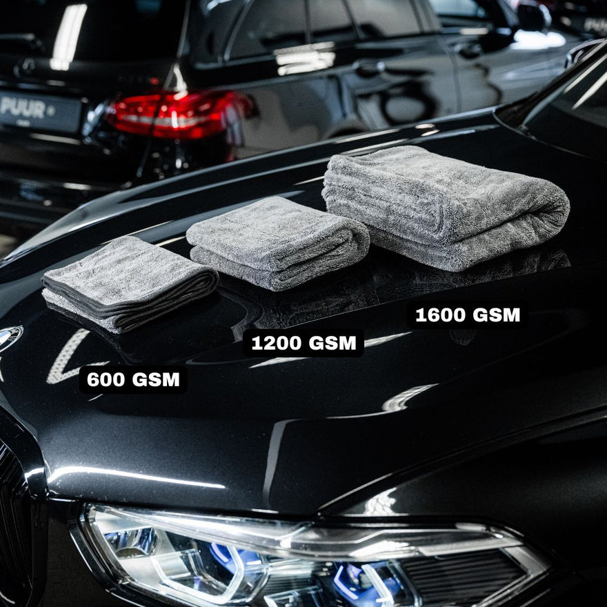 Three gray microfiber towels, including the Master Drying Towel 1600 GSM XL, are displayed on the hood of a shiny black car in a garage with other cars visible in the background.