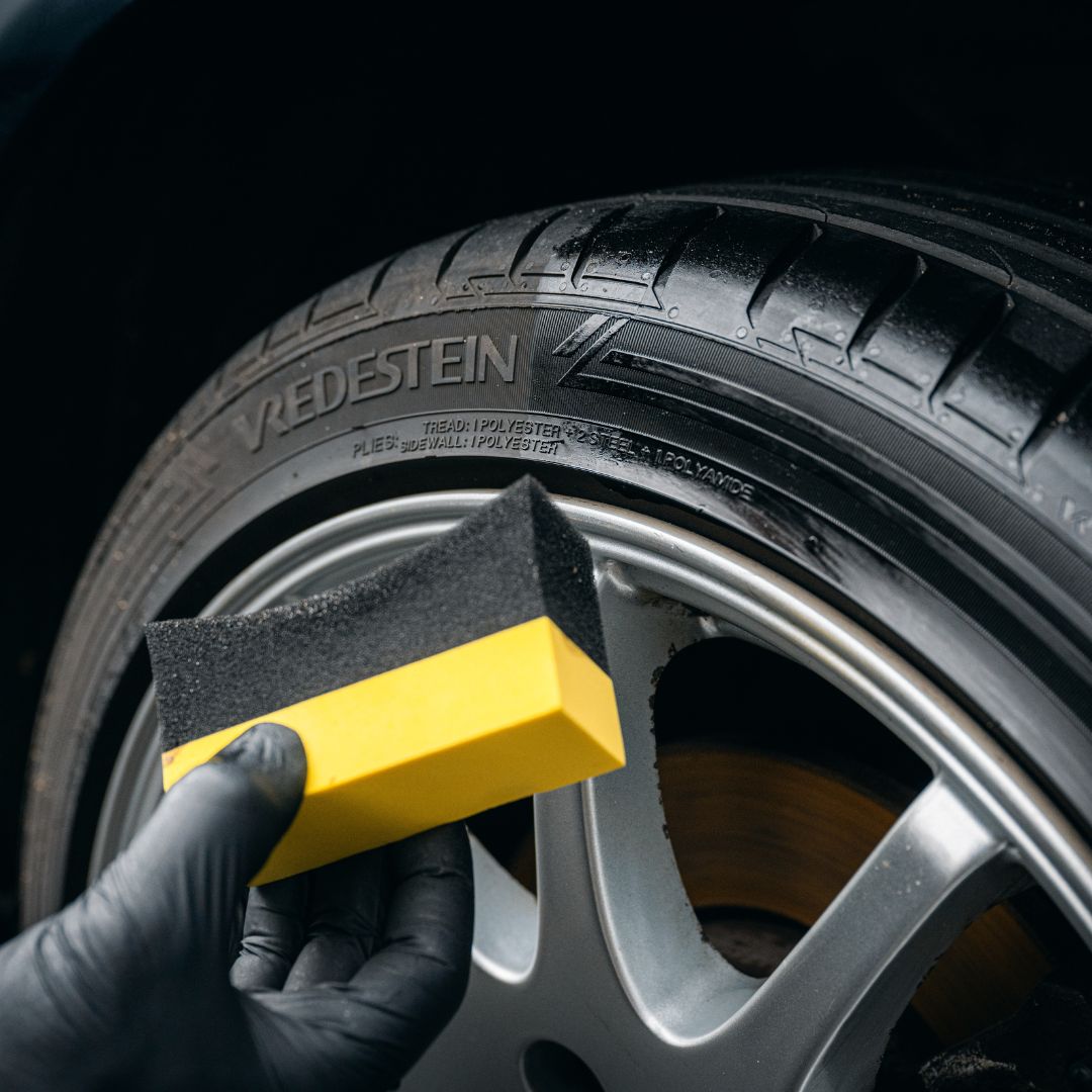 A gloved hand uses a yellow and black sponge to clean a black Vredestein tire on a silver alloy wheel—ideal for any car detailing routine with the Starter Kit | 21-delig.