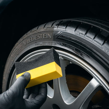 A gloved hand uses a yellow and black sponge to clean a black Vredestein tire on a silver alloy wheel—ideal for any car detailing routine with the Starter Kit | 21-delig.