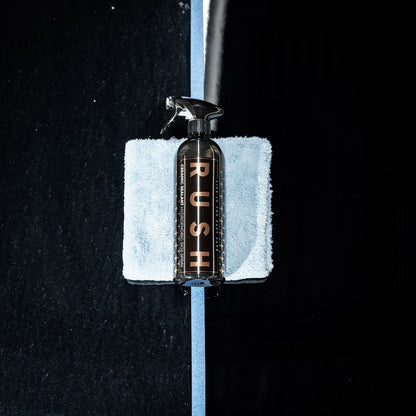 A spray bottle labeled Winter Protect DEAL, designed for winter protection, rests on a folded light blue microfiber cloth atop a reflective black surface divided by a strip of blue tape.