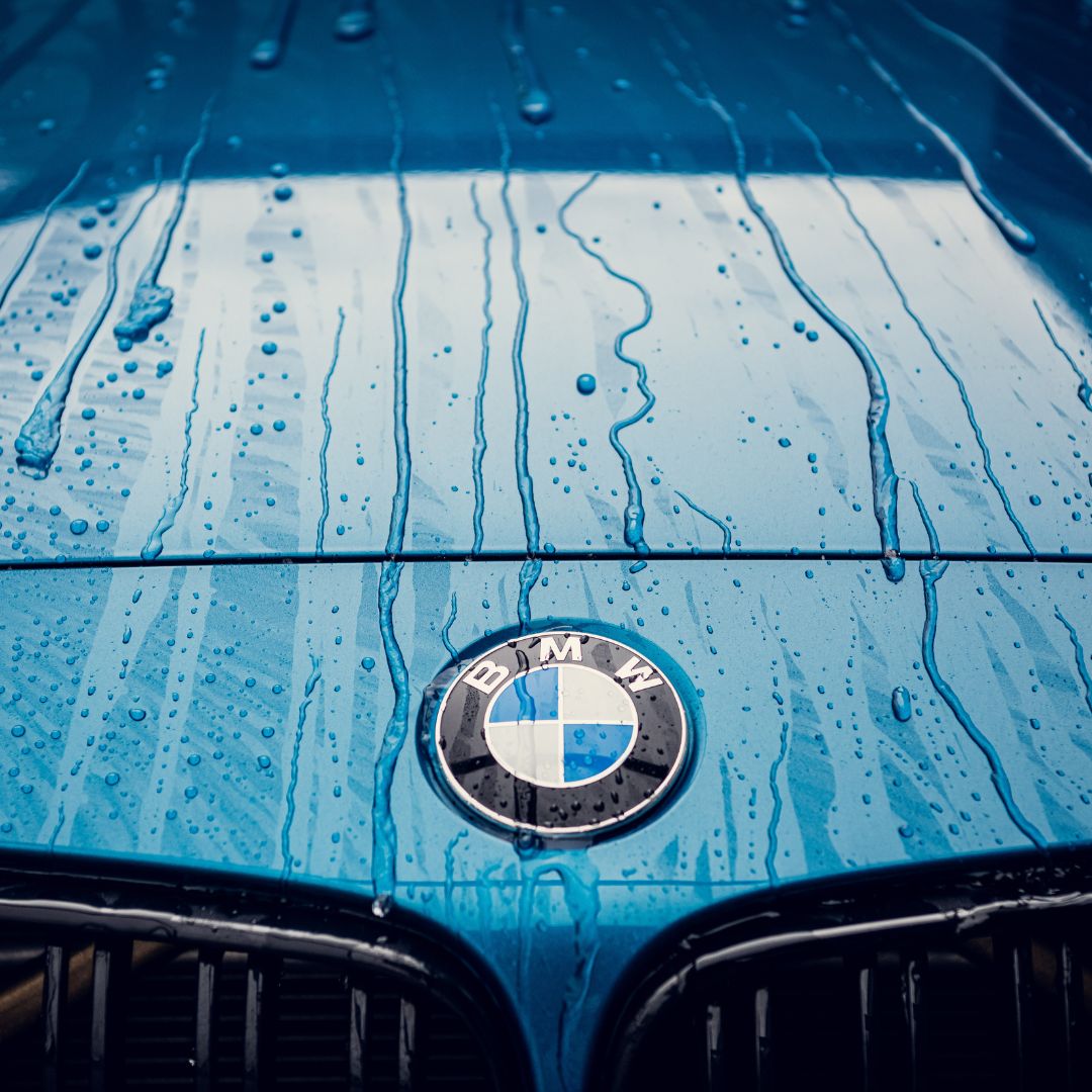 Close-up of a blue BMW hood with water droplets, focused on the BMW logo. RC Nano Wet Coat (TEST) creates een diepe glans and waterafstotend effect, making light reflect beautifully from above.