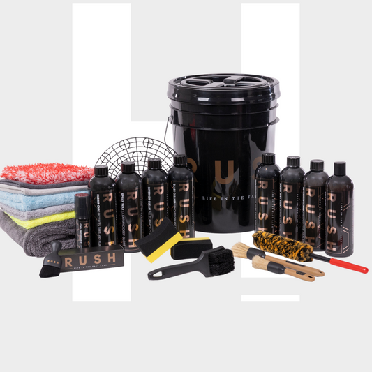 The Starter Kit XXL | 25-delig is a premium car cleaning set with black "RUSH" bottles, brushes, sponges, and microfiber towels, all neatly arranged around a large black bucket on a light gray background.