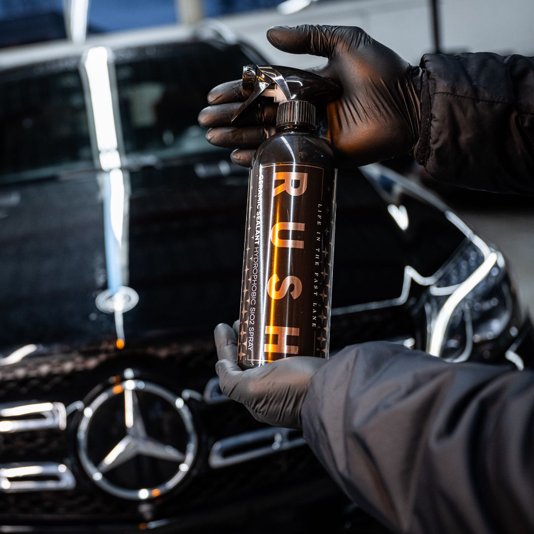 A person wearing black gloves holds a bottle labeled Ceramic Sealant Package SiO2 spray in front of a shiny black Mercedes-Benz, highlighting advanced car care and detailing.