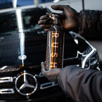 A person wearing black gloves holds a bottle labeled Ceramic Sealant Package SiO2 spray in front of a shiny black Mercedes-Benz, highlighting advanced car care and detailing.