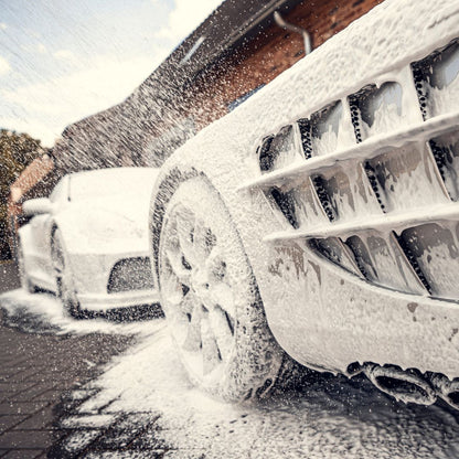 Close-up of a luxury sports car covered in a thick layer of Snow Foam during pre-wash, suds spraying as another car waits in the background. The outdoor scene takes place on a paved surface.