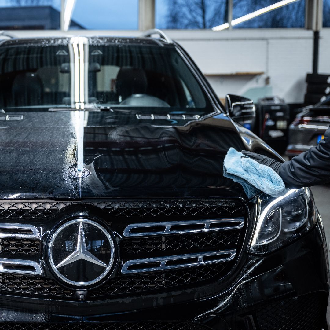 A person uses a blue microfiber cloth to wipe the hood of a shiny black Mercedes-Benz SUV in a garage, applying the Ceramic Sealant Package for extra paint protection.