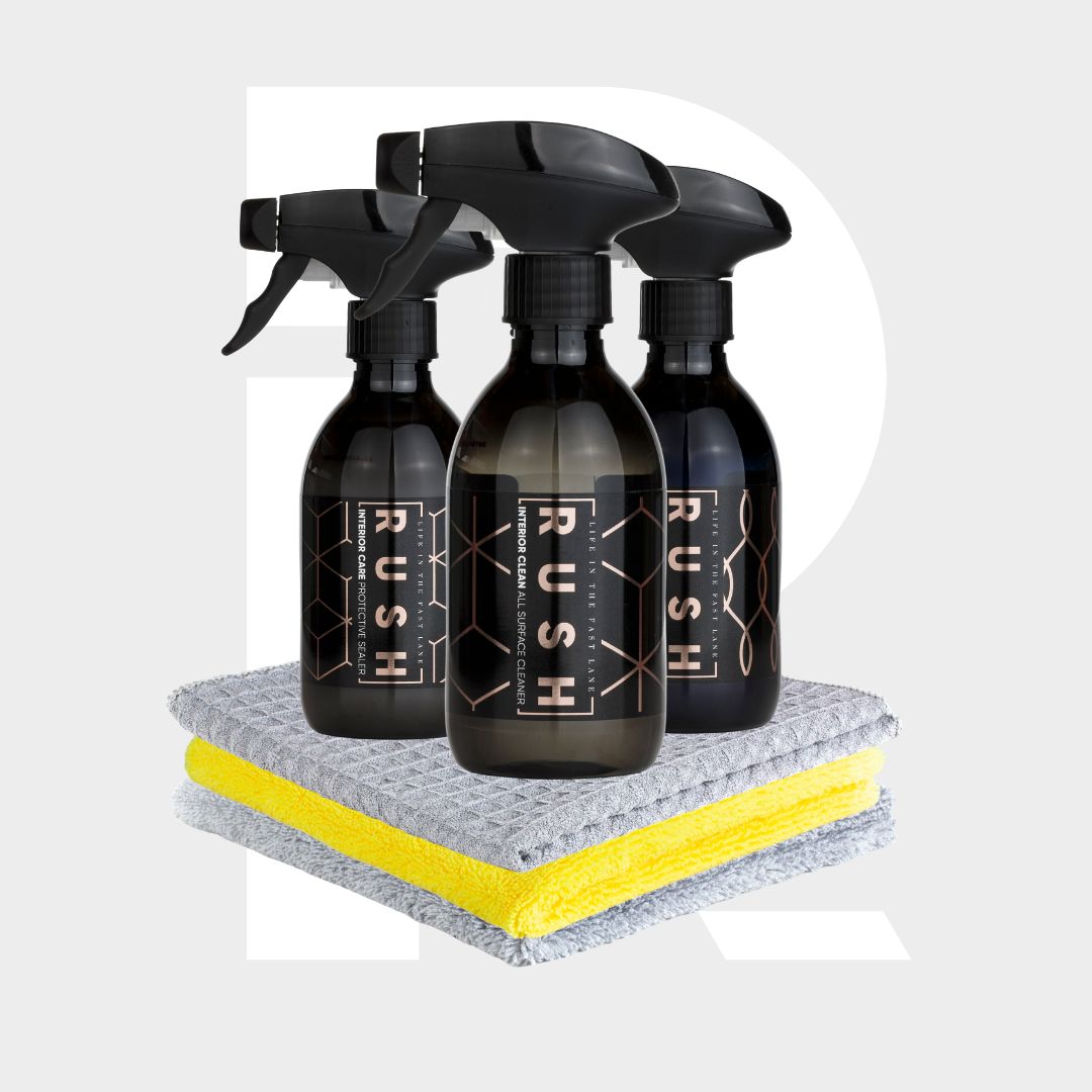 The Interior Sample Package includes three black RUSH spray bottles arranged behind two neatly folded microfiber cleaning cloths, one gray and one yellow, set against a light gray background with a large white "R".