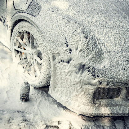 A car is coated in thick white Snow Foam during a pre-wash, covering the tire, wheel, and body. A small bottle of Snow Foam appears by the front wheel.