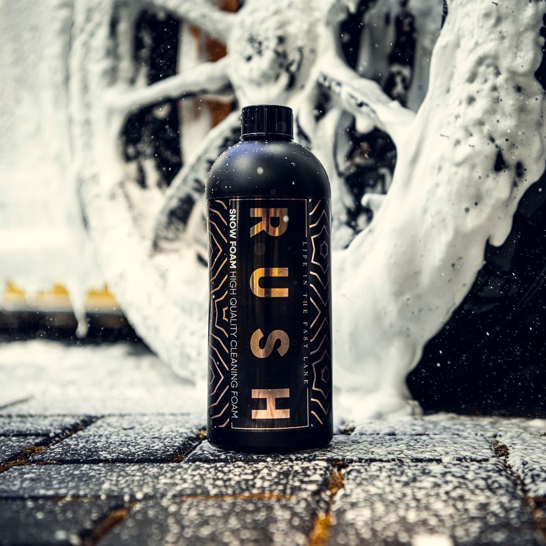 A black bottle labeled Snow Foam stands upright on wet, foamy ground, with a car wheel coated in thick white cleaning foam in the background. Snow Foam is scattered across the ground and surfaces.