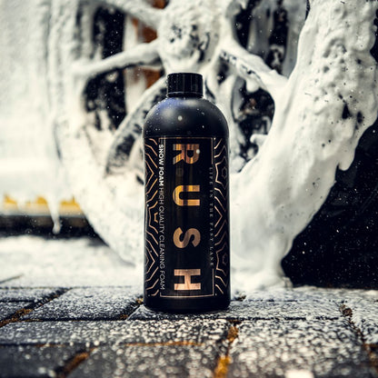 A black bottle labeled Snow Foam stands upright on wet, foamy ground, with a car wheel coated in thick white cleaning foam in the background. Snow Foam is scattered across the ground and surfaces.