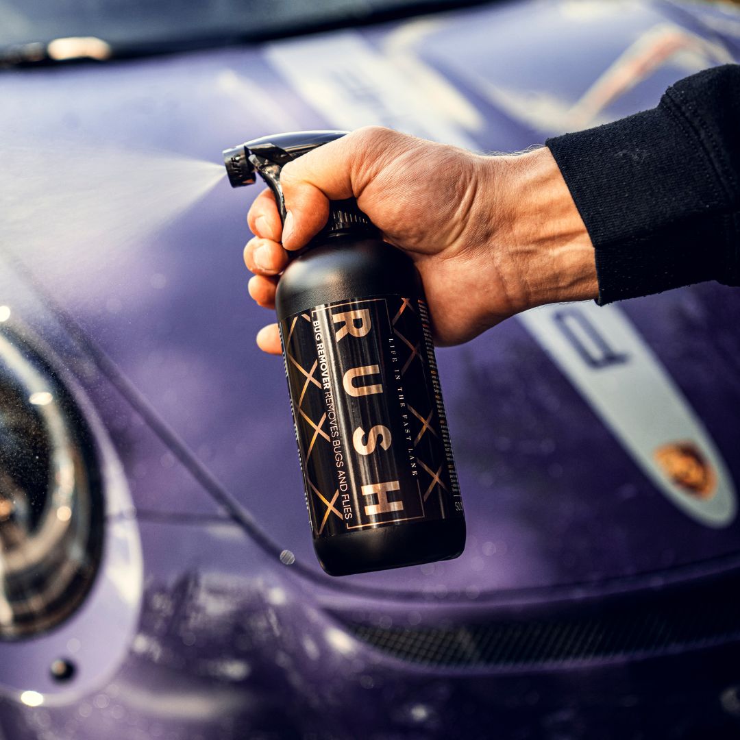 A hand holds a black spray bottle labeled RUSH in front of a purple Porsche, showcasing the exclusive Detailing Package PLUS | 23-delig auto reinigingspakket.