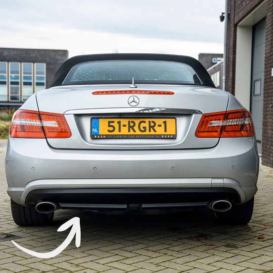 Rear view of a silver Mercedes-Benz convertible on a brick driveway with Dutch plate 51-RGR-1, arrow highlighting black kunststof exterieuronderdelen like the rear diffuser and dual exhaust pipes treated with Trim & Plastic Care.