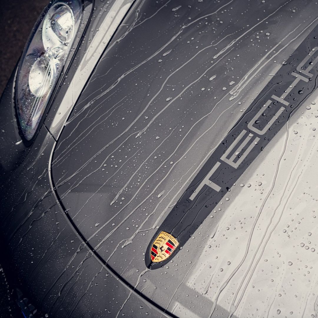 Close-up of a wet grey Porsche Taycan hood with raindrops, the Porsche emblem, and part of the headlight. The diepe glans and waterafstotend effect from RC Nano Wet Coat (TEST) enhance the car’s beauty, with 'Taycan' visible on its surface.