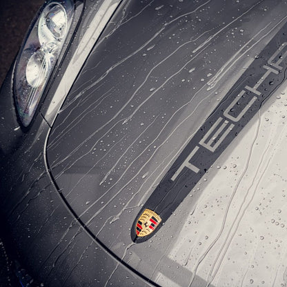 Close-up of a wet grey Porsche Taycan hood with raindrops, the Porsche emblem, and part of the headlight. The diepe glans and waterafstotend effect from RC Nano Wet Coat (TEST) enhance the car’s beauty, with 'Taycan' visible on its surface.
