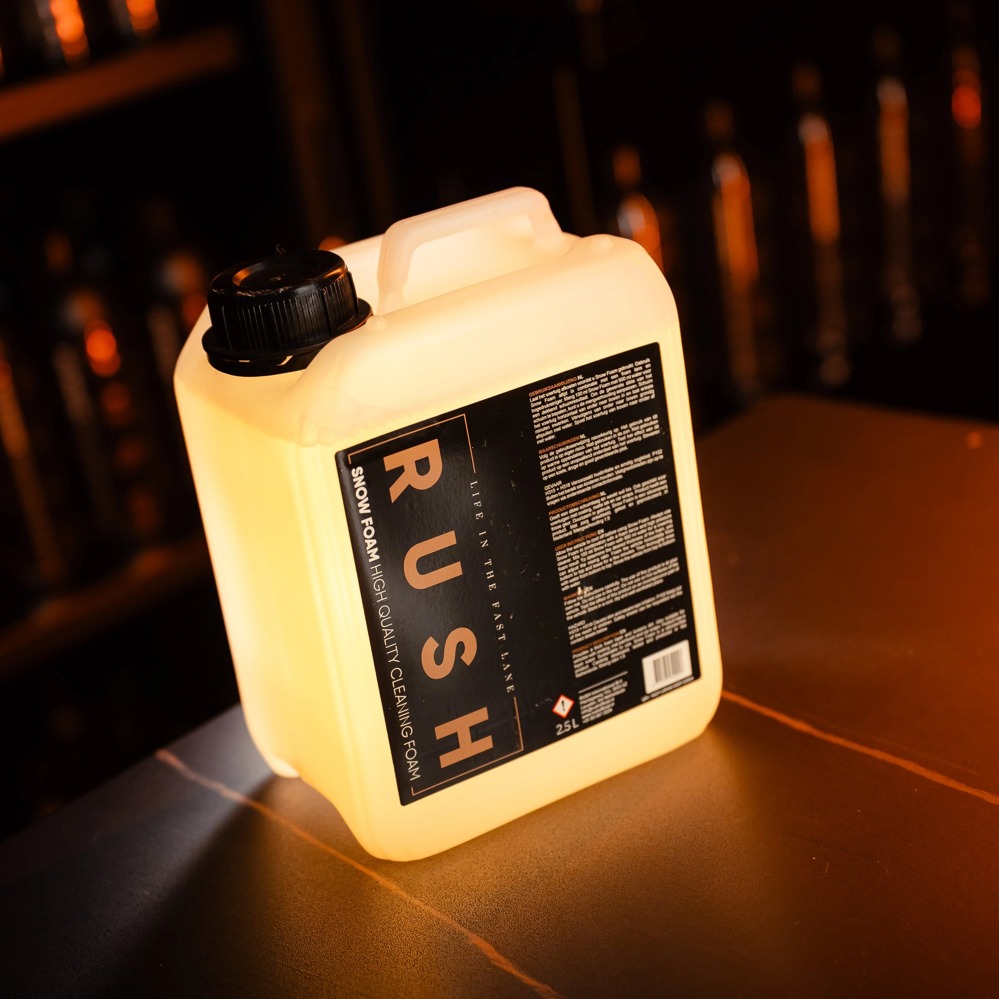 A translucent 2.5L plastic container labeled Snow Foam 2,5 liter rests on a countertop under warm lighting, with blurred bottles in the background.
